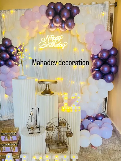 A chic home birthday decoration with a balloon arch in shades of purple and white. The setup is accented with a lit-up tree prop and a 'Happy Birthday' neon sign.