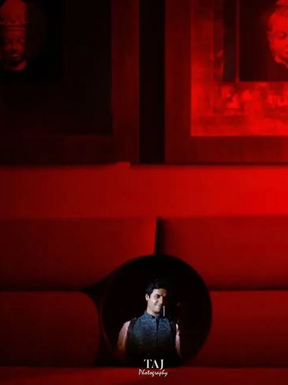 A creative framing shot, where the groom is seen through a circular opening in a vibrant red couch. This kind of composition is playful, artistic, and creates a visually interesting portrait.