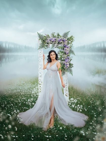 A full view of the ethereal setup by the lake. The combination of the flowing dress, floral arch, and misty atmosphere makes for a breathtaking maternity photograph.