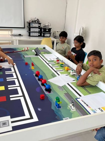 A group of students gathered around the WRO competition mat, strategically placing their completed game elements. This phase is crucial for understanding the challenge layout and planning their robot's mission path.