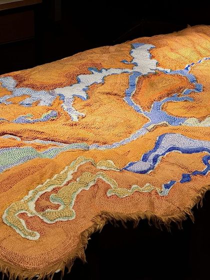 A textile map from 'Cu+Fibers', laid out for viewing. The piece uses different colors and stitching techniques to delineate land and water, transforming a map into a work of fiber art.