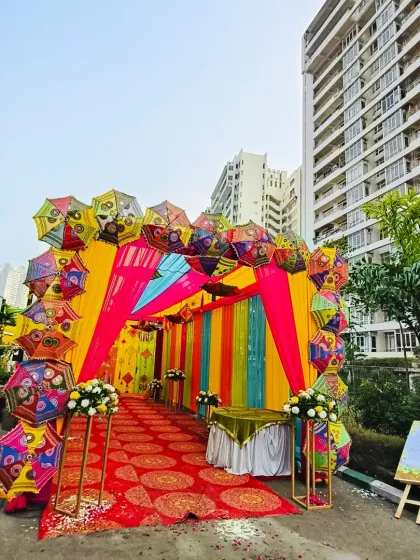 A grand and festive entrance to welcome your guests. This setup features a vibrant tunnel of pink, yellow, and blue drapes, framed by an arch made of colorful Rajasthani umbrellas, setting a joyful tone for a Mehendi or Haldi event.