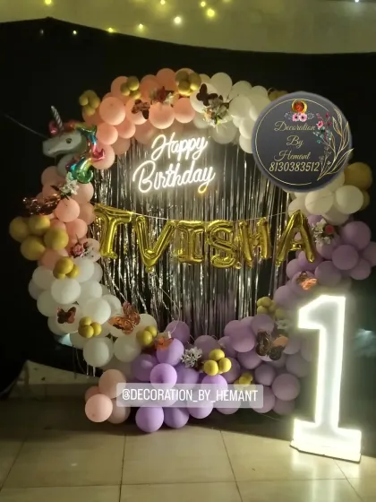 A unicorn-themed first birthday photo booth. The circular backdrop is framed with pastel pink, purple, and white balloons, a unicorn foil balloon, and a lit-up marquee number '1'.