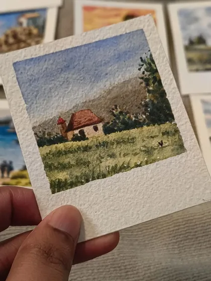This is a close-up of one of my favorite miniature watercolor Polaroids. I paint each tiny landscape by hand, capturing scenes like this rustic cottage nestled in a green field. The textured paper gives it an authentic, vintage feel.