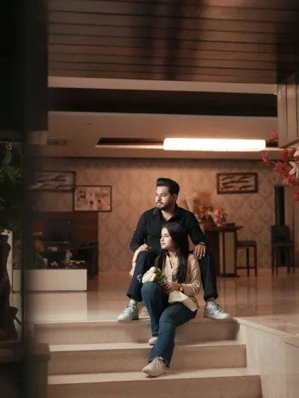 A full-length portrait of a couple sitting on the steps of a modern home, looking away together as if contemplating their future. The warm indoor lighting adds a cozy feel.