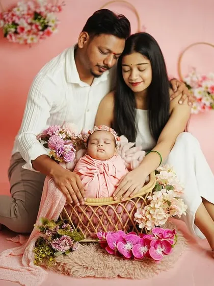 A serene family portrait with the parents and their sleeping newborn. The coordinated white outfits and pink floral theme create a harmonious and gentle image.