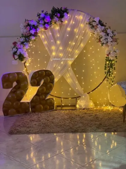 A simple yet beautiful backdrop for a 22nd birthday. The setup features a large ring frame decorated with fairy lights, sheer white drapes, and floral arrangements.