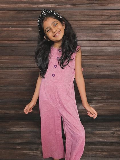 Another look at our chic pink cotton jumpsuit. It features pleat details, cute flower buttons, and a comfy fit with a back zipper. Perfect for playdates and parties!