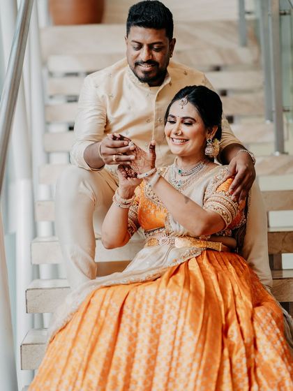 A couple seated on a staircase, sharing a sweet moment. Her orange and cream half saree and his cream kurta create a warm, coordinated look.