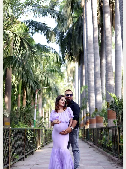 A full-length couple's portrait on a walkway surrounded by tall palm trees. The mother-to-be wears a lovely lavender mermaid-style gown.
