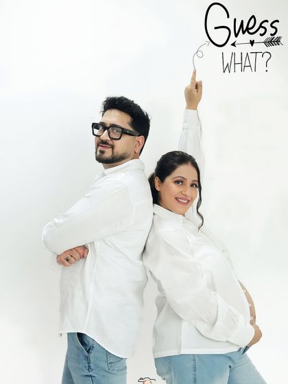 A playful "Guess What?" announcement photo for an expecting couple. The clean white background and matching outfits create a fresh and modern look for their baby announcement.