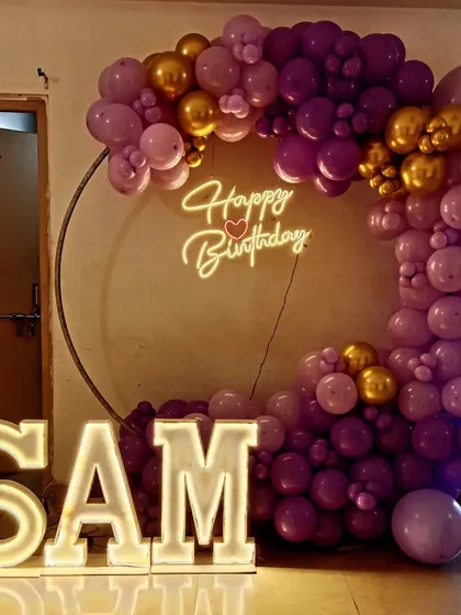 A personalized birthday setup with a purple and gold balloon ring. Large, illuminated letters spelling 'SAM' add a unique and personal touch to the decoration.