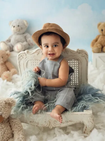 A baby boy in a cute hat sits on a vintage-style chair, surrounded by teddy bears and clouds. This setup creates a dreamy, whimsical feel.