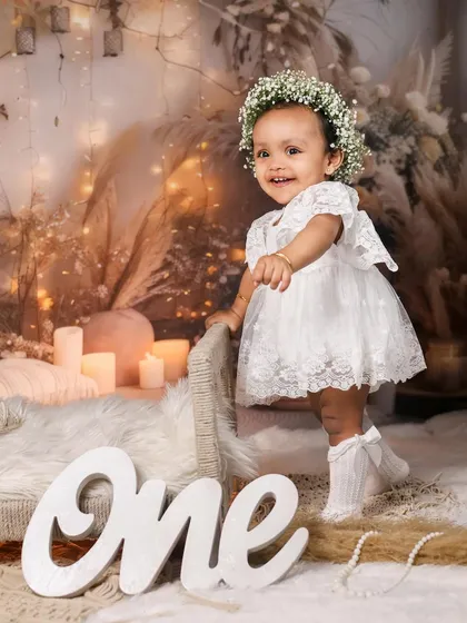 A smiling one-year-old girl in a white lace dress and floral crown, standing by a miniature bed in a boho-chic setup.