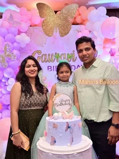 A happy family photo from a gorgeous butterfly-themed birthday. The backdrop features a shimmer panel, large butterfly wing props, and a soft pink balloon garland.
