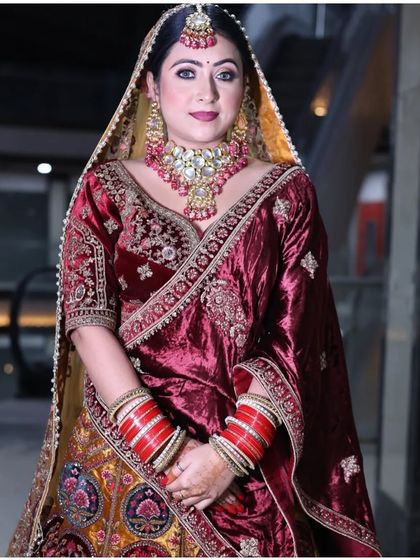 A full-length shot of a bride in a regal maroon and gold lehenga. This image displays the complete ensemble, including the heavy jewellery and traditional red chooda, perfect for a classic wedding look.