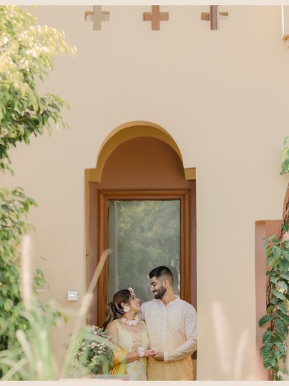 A quiet, romantic moment during the Haldi festivities. The couple holds hands, sharing a loving gaze away from the main celebration, framed by lush greenery.