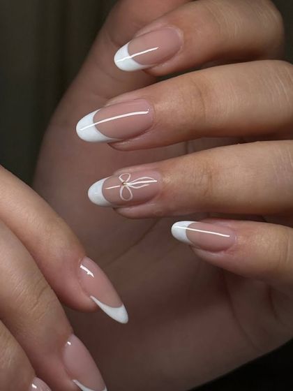 A delicate and sweet French manicure on almond-shaped nails. The classic white tips are complemented by a single, hand-painted white bow on the ring finger.