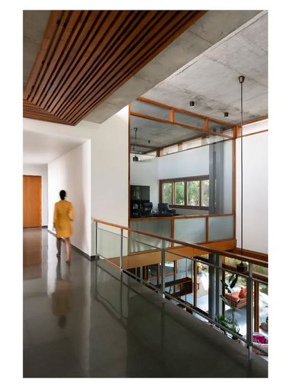 The upper corridor of the 'House of Canopies' features a slatted teak wood ceiling, which adds warmth and a linear rhythm that contrasts with the board-formed concrete of the walls.