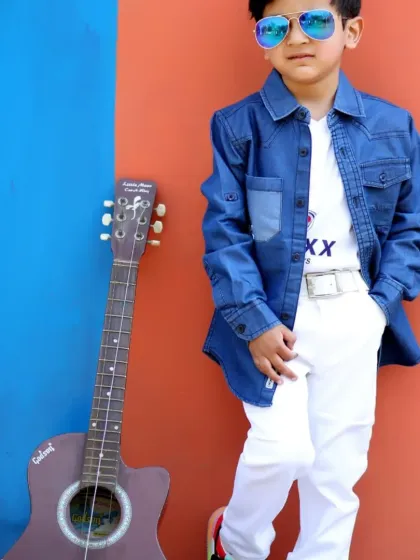 A young boy looking cool in a denim jacket and sunglasses, posing with a guitar against a colorful wall.