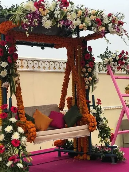 This Mehendi swing (jhoola) is heavily decorated with marigold garlands and other colorful flowers. The setup includes vibrant cushions and a pink decorative ladder, creating a perfect photo opportunity.