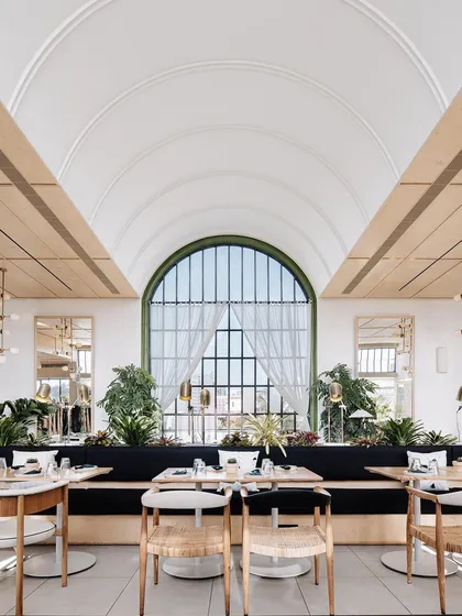 The main dining hall at Brassa is designed to feel bright, airy, and connected to the outdoors. The grand barrel-vaulted ceiling and large arched window create a sense of scale, while lush greenery and natural materials make the space feel welcoming and serene.