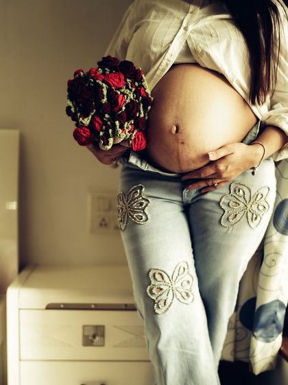 A stylish indoor shot focusing on the mom-to-be's fashion, pairing embroidered jeans with a bare bump and a bouquet of red roses.