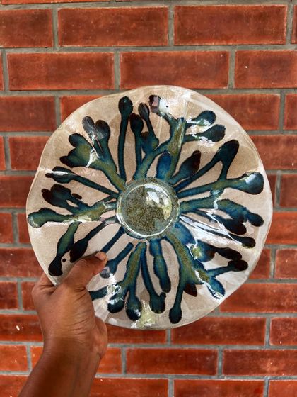 An abstract, flower-like bowl with a stunning blue and green glaze that bursts from the center. This piece was handmade by a student and shows a wonderful creative spirit.