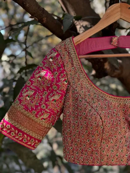 A close-up of a pink blouse featuring peacock motifs embroidered with silk thread and zardosi. The sleeve cuff has a thick, ornate border, adding to its grandeur.