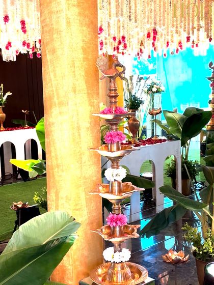 A tall, tiered brass lamp decorated with pink and white flowers, placed amidst lush green banana leaves, a classic element in traditional wedding decor.