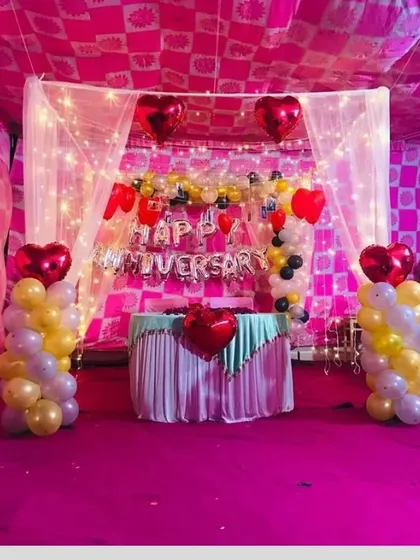 A classic anniversary stage setup under a tent. The decoration includes a 'Happy Anniversary' foil banner, heart-shaped balloons, and balloon columns in red, yellow, and white.