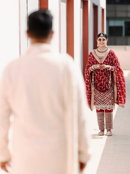 A "first look" style shot, with the groom out of focus in the foreground and the bride smiling in her beautiful red suit in the background. It builds a sense of anticipation and story.