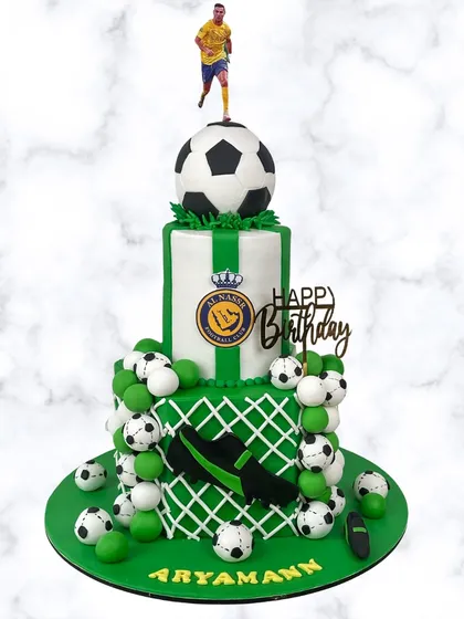 A grand, two-tiered soccer cake for Aryamann's birthday. This design includes the Al-Nassr logo, a goal net, cleats, and plenty of soccer balls for a sporty celebration.