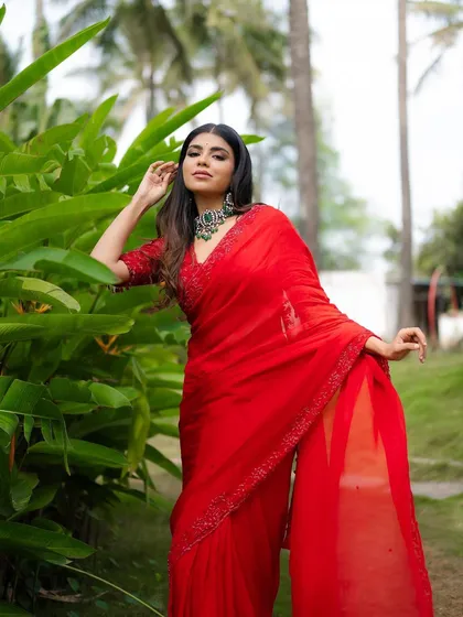 A full-length portrait of a model in a stunning red saree, standing amidst lush greenery. The composition highlights the elegance of the outfit in a natural setting.
