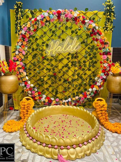 A joyful Haldi setup featuring a large circular floral ring against a leafy backdrop. The design is centered around a traditional brass urli, with golden pots adding a touch of elegance.