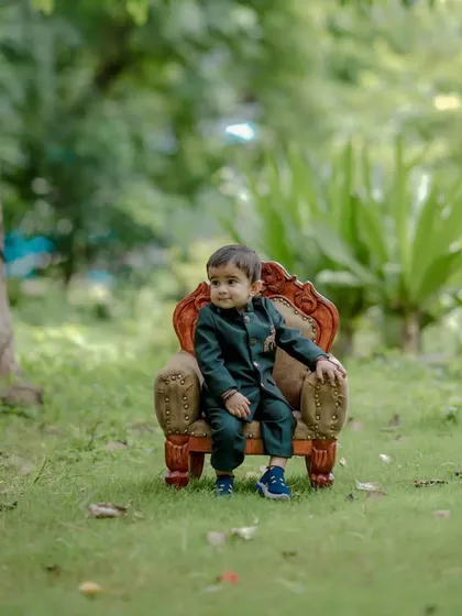 A wide shot showing the little boy in his smart suit, seated on the miniature throne in a beautiful park. This type of portrait captures both the child and the lovely outdoor environment of the shoot.