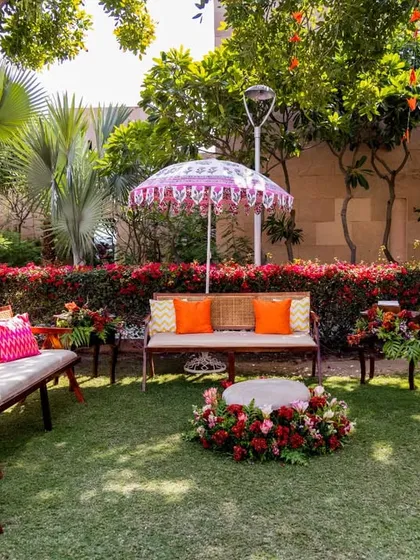 A comfortable and stylish lounge area for the Mehendi, set up on the lawn. The decor features wooden furniture, colorful pink and orange cushions, and a hand-painted parasol.