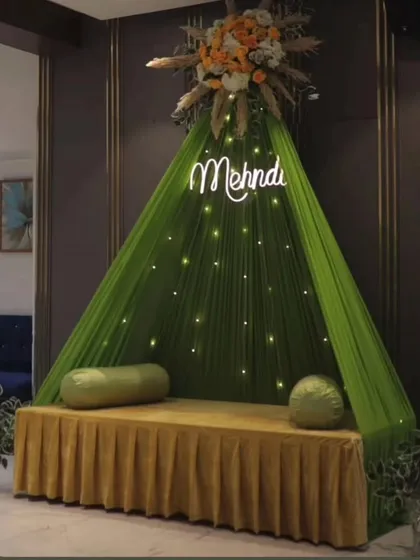 A modern and chic Mehandi corner. This design features a stylish teepee-style seating arrangement with green fabric, a custom "Mehndi" neon sign, and subtle fairy lights, perfect for contemporary brides who want a trendy photo-op.