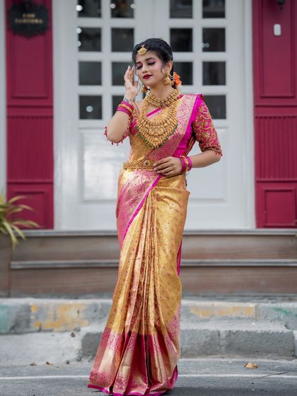 A full-length view of a stunning bridal look. The golden Kanjeevaram saree with a bright pink border is the centerpiece, complemented by heavy temple jewellery and makeup that makes the bride shine.
