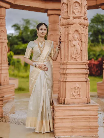 An elegant full-length portrait of a bride in a classic white and gold saree, standing beside intricately carved pillars that complement her grace.