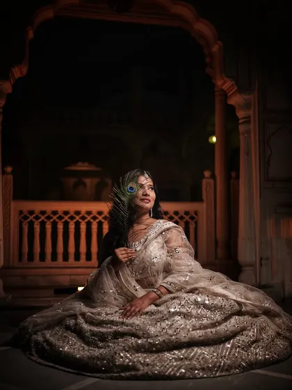 A bride in a sparkling lehenga sits in a beautifully lit heritage location, holding a peacock feather.