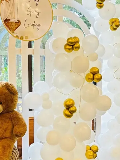 This close-up shows a white and gold balloon arch next to a seated teddy bear. The 'Baby is Landing' sign adds a cute, thematic touch to this hot-air balloon inspired decor.