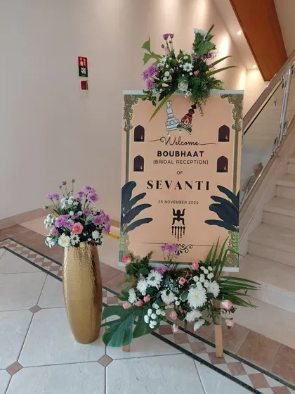 A custom welcome board for a Bengali Boubhaat, or bridal reception. The sign is decorated with culturally relevant motifs and surrounded by floral arrangements that match the event's color scheme.