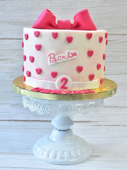 A chic and sweet Barbie-themed cake for a 2nd birthday. This cake is decorated with pink hearts, a large pink fondant bow on top, and the iconic Barbie logo.
