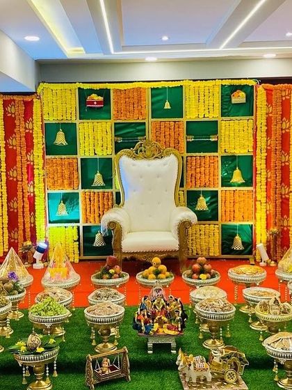 A full view of a traditional Seemantham stage, complete with a floral checkerboard backdrop and an elaborate display of offerings. The setup includes traditional dolls and cow motifs for a truly authentic feel.