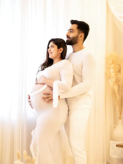 An elegant all-white themed couple portrait. The father-to-be stands behind, protectively holding the mother and her bump as they look towards the light.