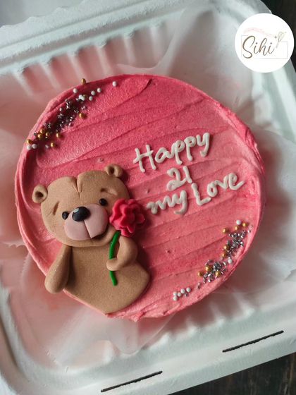A sweet teddy bear bento cake perfect for a loved one. The fondant bear is holding a single red rose, set against a textured pink buttercream background with sparkling sprinkles.