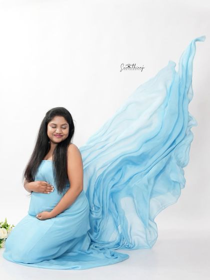 A dreamy portrait of a mother-to-be in a light blue gown with a flowing fabric effect. The simple white background creates an ethereal and angelic feel.