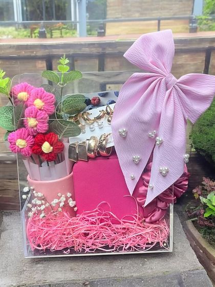 A bright and cheerful gift hamper for her, arranged in a clear acrylic box. This pink-themed set includes hair accessories, a large bow, and other cute items, perfect for a vibrant personality.