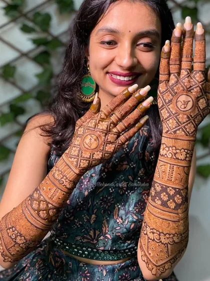 A happy bride showing off her intricate back-of-hand mehendi. The design features personalized initials within a classic grid pattern.